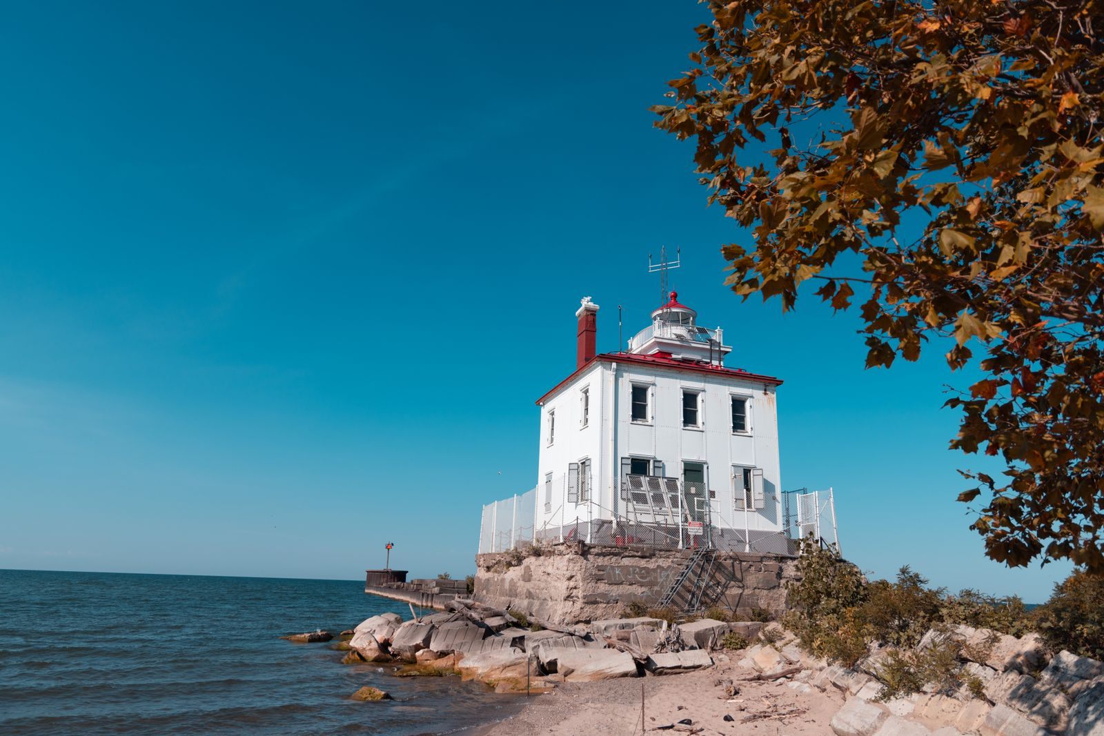 A white lighthouse with a red lantern on a rocky Lake Erie shore, framed by autumn leaves