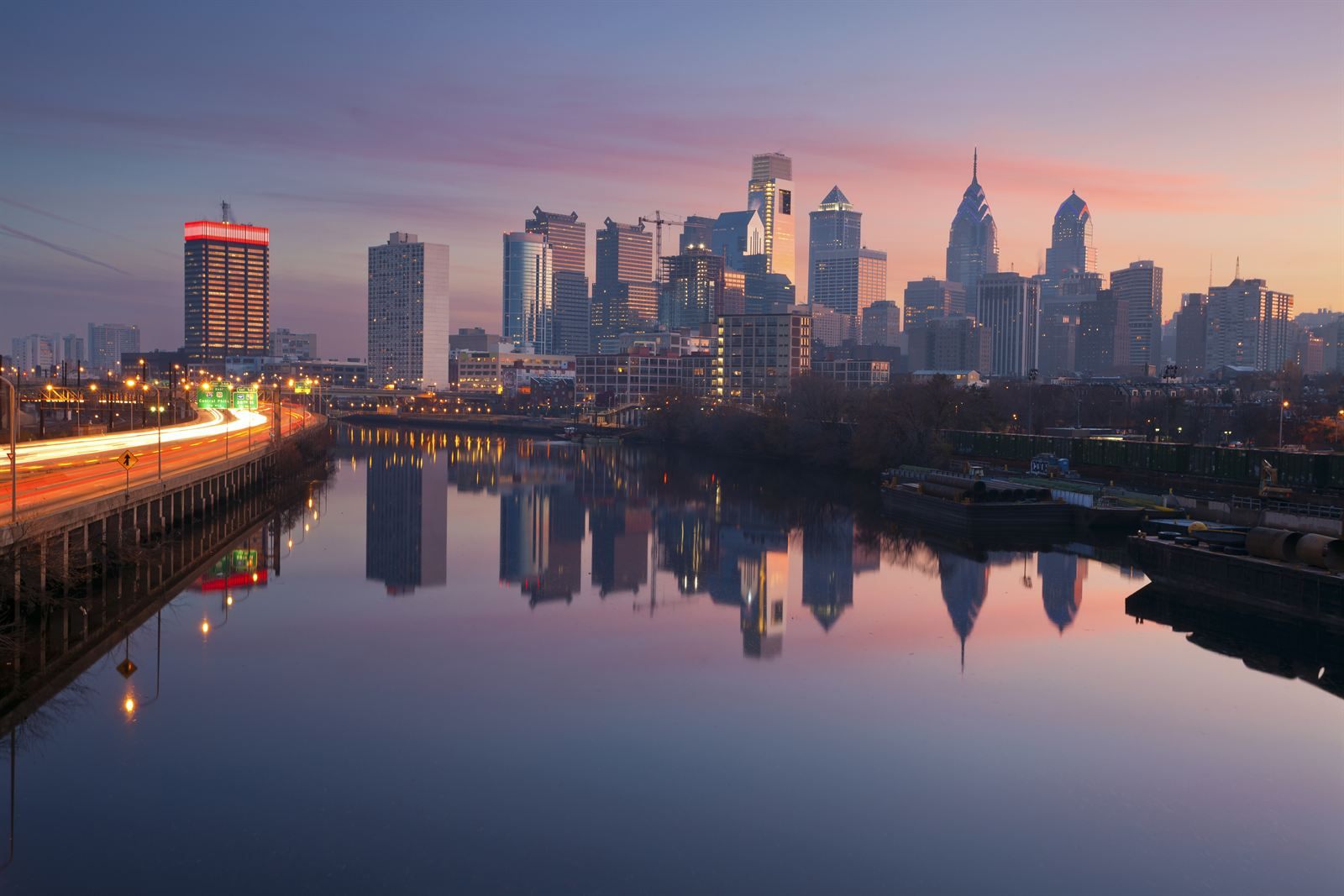 The Philadelphia skyline reflected in the Schuylkill River at dusk, with traffic light trails along the riverbank